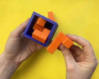 Può includere: Un contenitore quadrato blu che ospita diversi pezzi di puzzle arancioni composti da cubi, con un ulteriore pezzo di puzzle arancione a forma di L tenuto separatamente. I componenti del puzzle sono solidi blocchi arancioni.