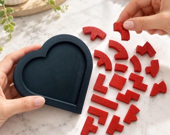 Può includere: Un vassoio nero a forma di cuore e una collezione di piccoli pezzi di puzzle geometrici rossi e testurizzati. I pezzi sono sparsi a destra del vassoio, con un pezzo tenuto sopra gli altri, pronto per essere inserito nella cornice vuota.