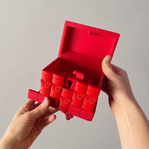 May include: A bright red, rectangular box with a hinged lid, featuring four vertical panels on the front, each embossed with a butterfly design. The box includes a central keyhole mechanism and a small, matching red key hanging from the bottom.