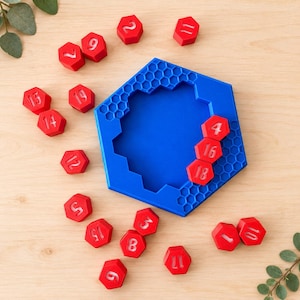 Könnte beinhalten: Ein blaues, sechseckiges Spielbrett mit Wabenmuster und einer großen Aussparung in der Mitte, begleitet von mehreren kleinen roten, sechseckigen Spielsteinen, die jeweils eine weiße Zahl von 1 bis 19 tragen, wobei drei Steine im Brett liegen.