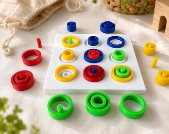Può includere: Una tavola quadrata bianca con una griglia tre per tre di rientranze circolari, dotata di vari anelli impilabili e pioli in colori primari tra cui rosso, blu, giallo e verde, con diversi pezzi sparsi attorno alla base.