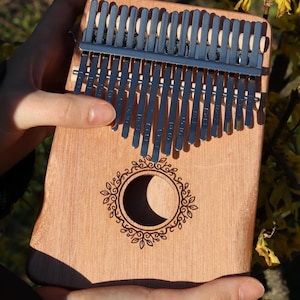 May include: A wooden kalimba featuring seventeen metal tines and a central circular sound hole. The sound hole is surrounded by an engraved leaf-patterned border. The instrument has a natural light brown wood grain finish and silver metal keys.