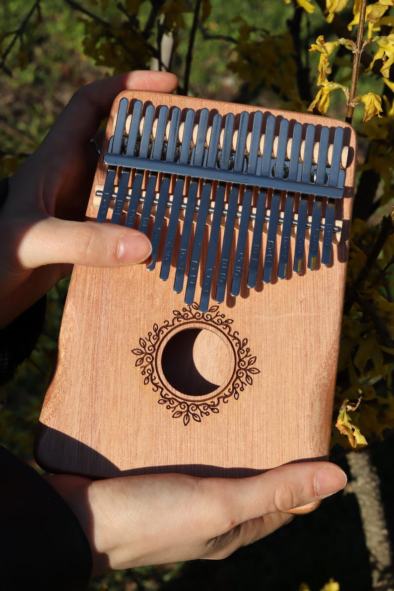 May include: A wooden kalimba featuring seventeen metal tines and a central circular sound hole. The sound hole is surrounded by an engraved leaf-patterned border. The instrument has a natural light brown wood grain finish and silver metal keys.