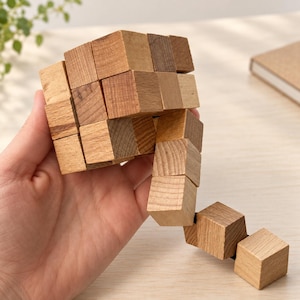 May include: A wooden puzzle consisting of multiple small, light brown cubes connected by an internal cord. The cubes are arranged in a partially collapsed, flexible structure with visible wood grain patterns on each individual block surface.