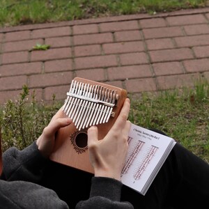 May include: A wooden kalimba with seventeen silver metal tines and a circular sound hole featuring a decorative black floral border. The instrument is held alongside an open instructional booklet displaying musical notation for Twinkle Twinkle Little Star.
