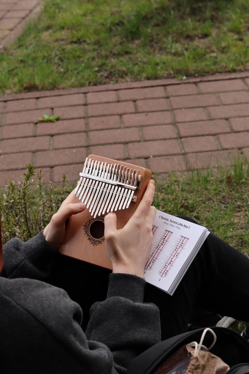 May include: A wooden kalimba with seventeen silver metal tines and a circular sound hole featuring a decorative black floral border. The instrument is held alongside an open instructional booklet displaying musical notation for Twinkle Twinkle Little Star.