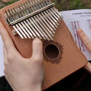 May include: A wooden kalimba with seventeen silver metal tines, each engraved with musical notes and numbers. The instrument features a central circular sound hole with a decorative leaf-patterned border and is accompanied by a music sheet.