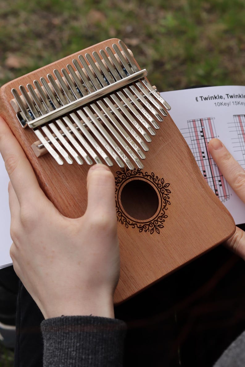 May include: A wooden kalimba with seventeen silver metal tines, each engraved with musical notes and numbers. The instrument features a central circular sound hole with a decorative leaf-patterned border and is accompanied by a music sheet.