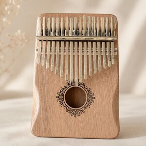 May include: A wooden kalimba with seventeen silver metal tines, each engraved with musical notes. The instrument features a central circular sound hole surrounded by a delicate, dark-etched floral vine pattern on the smooth, light brown wood body.