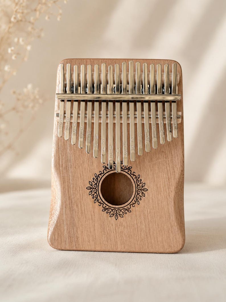 May include: A wooden kalimba with seventeen silver metal tines, each engraved with musical notes. The instrument features a central circular sound hole surrounded by a delicate, dark-etched floral vine pattern on the smooth, light brown wood body.
