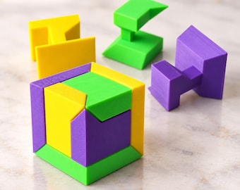 Può includere: Un cubo puzzle 3D multicolore assemblato con pezzi geometrici a incastro di colore giallo brillante, verde lime e viola. Tre ulteriori componenti del puzzle, negli stessi colori, sono posizionati separatamente vicino al cubo principale.
