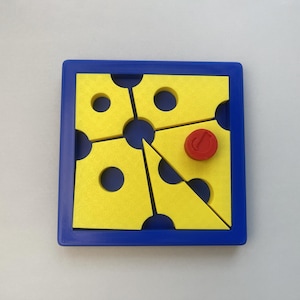 May include: A square blue frame containing four yellow puzzle pieces with circular cutouts. The pieces are arranged to form a central circular opening. A small red circular token with a musical note symbol is placed on one of the yellow pieces.