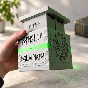 Könnte beinhalten: Eine graue, würfelförmige Box mit flachem Deckel und einem grünen horizontalen Band. Die Vorderseite zeigt den Text FHTAGN, PH'NGLUI und MGLW'NAFH in schwarzer Schrift. Die Seitenwand zeigt ein kreisförmiges Relief eines Tentakelwesens.