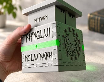 Può includere: Una scatola cubica grigia con una parte superiore piatta e una fascia orizzontale verde. La parte anteriore presenta il testo FHTAGN, PH'NGLUI e MGLW'NAFH in lettere nere. Il pannello laterale mostra un rilievo circolare di una creatura.