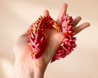 Può includere: Una statuetta di drago articolata e flessibile caratterizzata da una sfumatura di colore che passa dal rosso intenso all'arancione brillante e al giallo. La creatura presenta una superficie testurizzata e squamata con punte prominenti sul dorso.