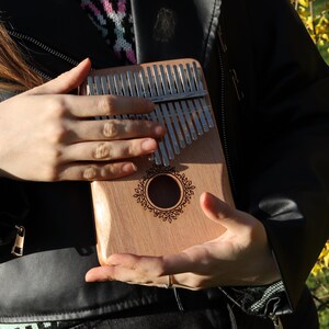 May include: A wooden kalimba featuring a series of silver metal tines arranged in a row across the top. The instrument has a central circular sound hole surrounded by a decorative engraved floral pattern on the light brown wooden body.