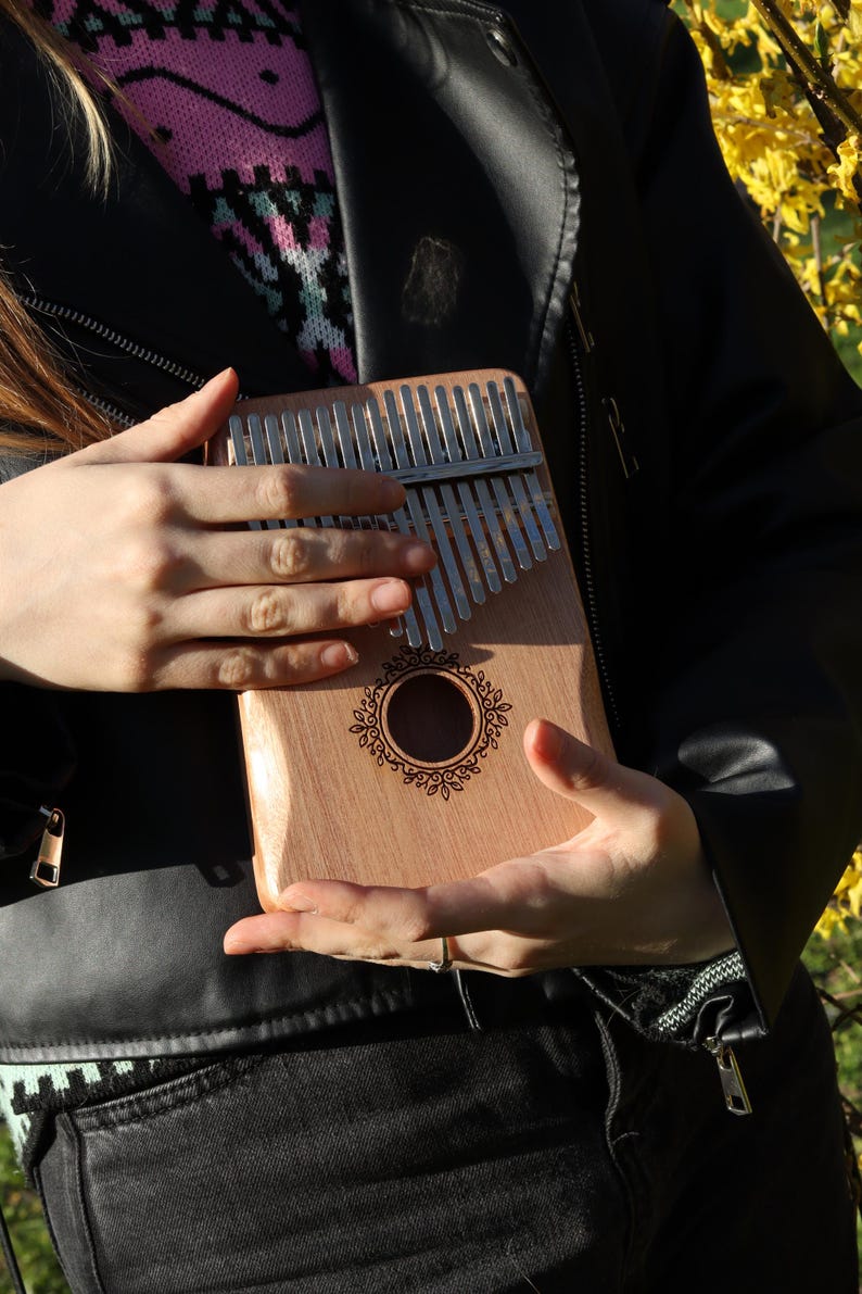 May include: A wooden kalimba featuring a series of silver metal tines arranged in a row across the top. The instrument has a central circular sound hole surrounded by a decorative engraved floral pattern on the light brown wooden body.