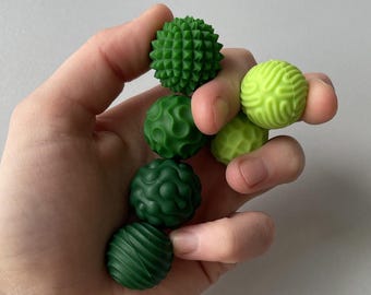 May include: A collection of five small, spherical sensory balls in various shades of green and lime. Each ball features a distinct tactile surface texture, including spikes, swirling patterns, and ridges, designed for sensory stimulation and tactile play.