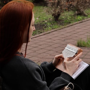 May include: A wooden kalimba with silver metal tines and a circular decorative engraving on the front. The instrument is held in both hands, accompanied by a small white instructional booklet with musical notation held alongside the wooden body.