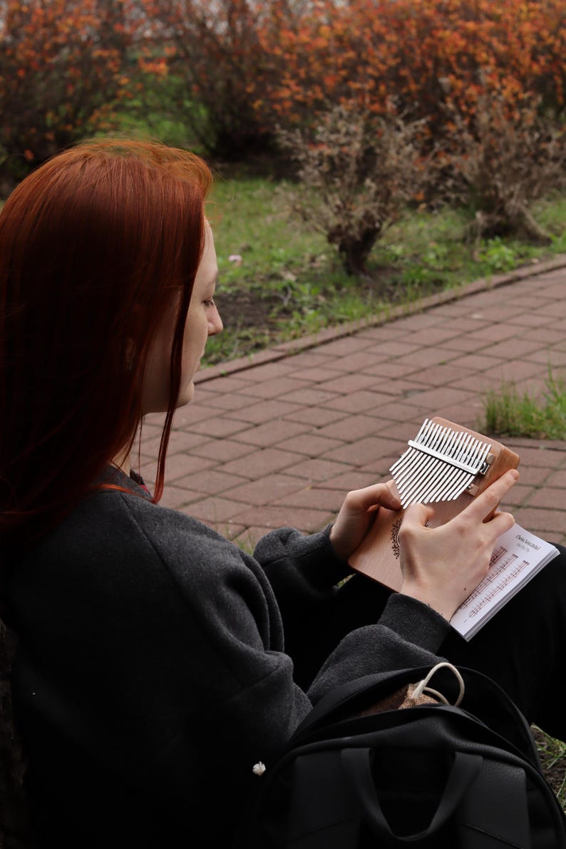 May include: A wooden kalimba with silver metal tines and a circular decorative engraving on the front. The instrument is held in both hands, accompanied by a small white instructional booklet with musical notation held alongside the wooden body.