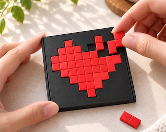 Può includere: Una tavola da puzzle quadrata nera con un design a cuore pixelato rosso. La tavola presenta spazi a griglia incassati dove vengono posizionate piccole tessere quadrate rosse per formare il cuore, con una tessera tenuta in mano.