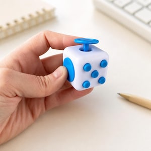 Può includere: Un cubo antistress bianco con elementi tattili blu, tra cui un pulsante superiore circolare, un grande pulsante laterale e un gruppo di cinque pulsanti circolari più piccoli sulla parte anteriore, progettato per la stimolazione sensoriale.
