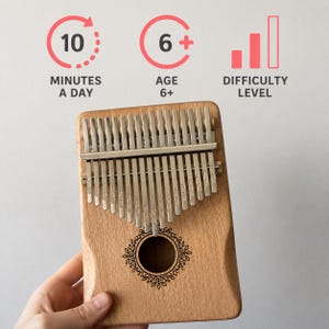 May include: A wooden kalimba with seventeen metal tines featuring engraved musical notes. Above the instrument, text and icons indicate 10 MINUTES A DAY, AGE 6+, and DIFFICULTY LEVEL. The kalimba has a central circular sound hole with a decorative border.