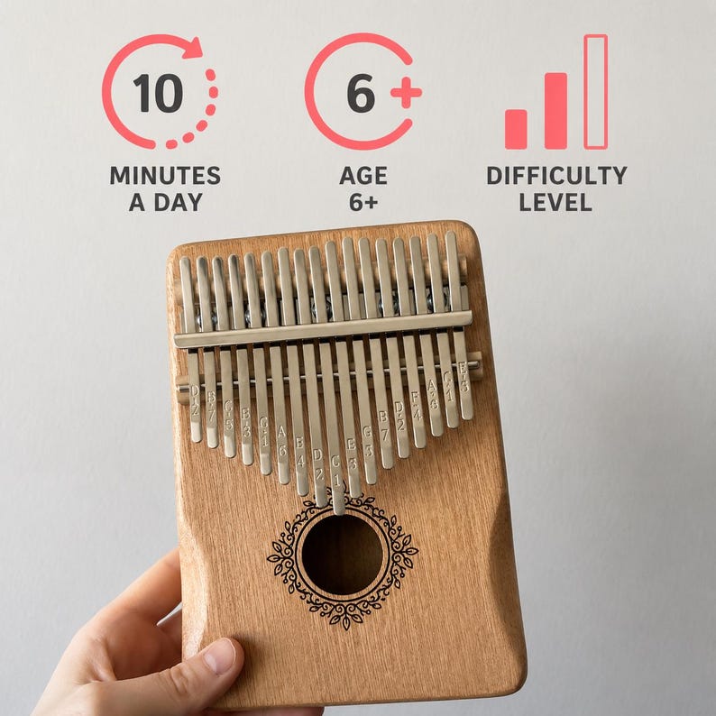 May include: A wooden kalimba with seventeen metal tines featuring engraved musical notes. Above the instrument, text and icons indicate 10 MINUTES A DAY, AGE 6+, and DIFFICULTY LEVEL. The kalimba has a central circular sound hole with a decorative border.