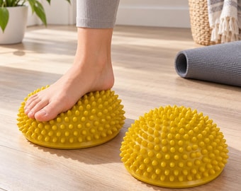 May include: Two yellow, dome-shaped balance pods featuring a textured surface covered in numerous small, rounded spikes. One pod is positioned on the floor, while the other is being stepped on by a bare foot to demonstrate its functional use.