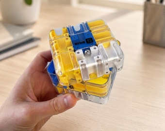 Può includere: Un cubo rompicapo in plastica trasparente con percorsi interni gialli, bianchi e blu e una piccola sfera di metallo. Il pezzo quadrato centrale blu riporta la scritta MAZE. Il rompicapo è composto da diversi segmenti interconnessi.