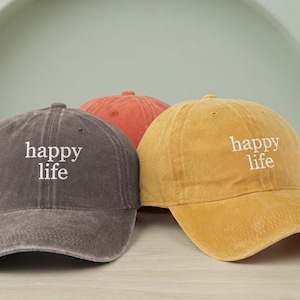 Pode incluir: Três bonés de beisebol nas cores cinza-carvão, laranja queimado e amarelo mostarda. Cada boné apresenta uma aba curva, um botão no topo e o texto bordado em branco "happy life" centralizado no painel frontal dos bonés cinza e amarelo.