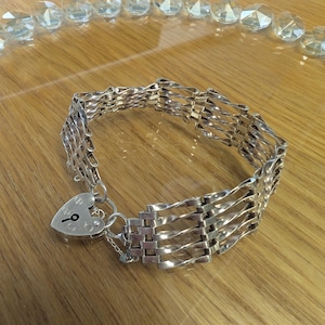 May include: A silver-toned bracelet featuring a multi-strand woven metal link design. The bracelet is secured with a heart-shaped padlock charm that includes a small keyhole detail on its front surface, connected by a circular jump ring.