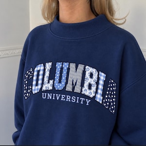 May include: A navy blue sweatshirt with a mock neck collar. The front features the word COLUMBIA in large, appliqued letters, each with a unique blue and white pattern including gingham, stripes, polka dots, and florals. Below, UNIVERSITY is printed.