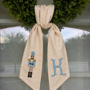 Könnte beinhalten: Ein cremefarbenes Stoffband mit Knoten, das auf dem linken Ende eine gestickte hellblaue Nussknacker-Figur und auf dem rechten Ende einen großen, hellblau gestickten Buchstaben K zeigt, mit glatter Stofftextur und sauberen Nähten.
