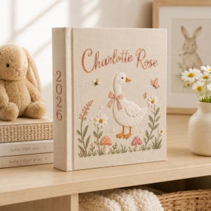Könnte beinhalten: Ein cremefarbenes, stoffbezogenes Fotoalbum mit gestickten Details einer Gans mit rosa Schleife, Blumen, Pilzen, einer Biene und einem Schmetterling. Der Name Charlotte Rose ist in rosa Schreibschrift gestickt und das Jahr 2026 steht auf dem Buchrücken.