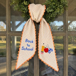 Könnte beinhalten: Ein Türhänger aus beigefarbenem und weißem Vichy-Stoff mit orangefarbenen, gewellten Rändern. Die linke Seite zeigt den blauen Sticktext Back to School. Die rechte Seite zeigt gestickte Symbole eines gelben Bleistifts, eines roten Apfels und einer blauen Schere.