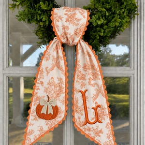 Könnte beinhalten: Eine lange, cremefarbene Stoffschärpe mit orangefarbenem Blumenmuster im Toile-Stil und gewellten Rändern. Eine Seite zeigt einen gestickten orangefarbenen Kürbis mit cremefarbener Schleife, die andere Seite einen großen, gestickten Buchstaben L.