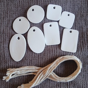 Peut inclure: Un ensemble de sept étiquettes en céramique blanche lisse de formes variées, incluant des cercles, des ovales et des rectangles, chacune dotée d'un trou pré-percé en haut. Une longue cordelette en coton beige naturel est incluse en dessous.