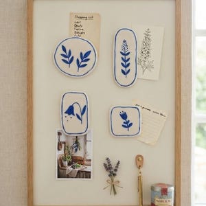 Peut inclure: Un tableau d'affichage rectangulaire à cadre en bois avec quatre aimants en céramique blanche ornés d'illustrations botaniques bleues, une liste de courses, un croquis floral, une photo, un brin de lavande séchée, un crochet en laiton et une boîte.
