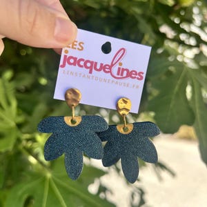 Könnte beinhalten: Ein Paar dunkelblaue, fächerförmige Ohrringe mit strukturierter Oberfläche und goldfarbenen Akzenten. Die Ohrringe sind an einer weißen Karte befestigt, die den Text "les jacquelines" und "linstantdunepause.fr" in rosa und orangefarbener Schrift zeigt.