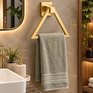 May include: A triangular gold-toned metal towel ring mounted on a wall, holding a folded sage green cotton hand towel with a textured border detail. The hardware features a square base plate and a geometric frame design for hanging bathroom linens.