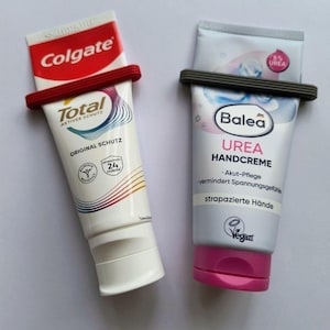 Könnte beinhalten: Zwei Tuben mit Kunststoffclips. Die linke Tube ist weiß und rot mit Colgate Total Branding. Die rechte Tube ist hellblau und weiß mit Balea Urea Handcreme Branding, einem rosa Verschluss und einem 5% Urea Etikett auf der Vorderseite.