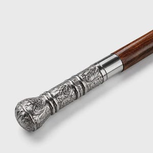 Può includere: Un bastone da passeggio con un fusto in legno marrone scuro e un'impugnatura in metallo color argento. L'impugnatura è cilindrica con una sommità arrotondata ed è decorata con intricati motivi floreali e a volute in rilievo sulla superficie.