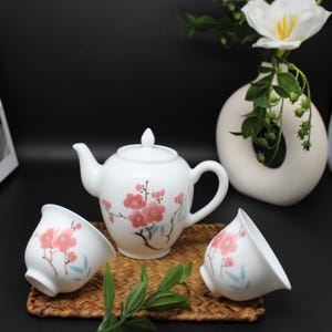 May include: A white porcelain tea set consisting of a teapot and two matching tea cups. Each piece features a delicate hand-painted design of pink plum blossoms with brown branches and light blue leaves against a smooth white surface.
