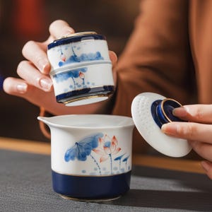 Puede incluir: Una taza de té de cerámica blanca con base azul profundo y borde dorado, con un diseño de flor de loto pintado a mano en azul y naranja. El juego incluye un filtro de té extraíble a juego y una tapa blanca con un pomo dorado.