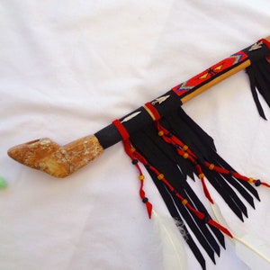 May include: A wooden pipe with black, red, and yellow beading and feathers. The pipe is decorated with black and white fringe.
