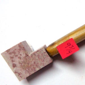 May include: A wooden-handled tool with a rectangular, speckled, pinkish-brown stone head. A small red tag with the number "93/4" is attached. The handle is a light brown color.