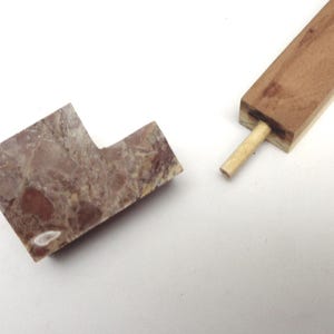 Può includere: Una pipa da tabacco smontata. La testa è in pietra marmorizzata marrone-rossastra. Lo stelo è un pezzo di legno rettangolare con un tassello cilindrico inserito. La pipa è mostrata su sfondo bianco.