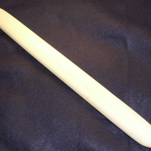 Peut inclure: Un rouleau à pâtisserie en bois, de couleur blanc cassé, de forme cylindrique et allongée. Il présente une extrémité arrondie et une petite cheville en bois à l'autre extrémité. Le fond est un tissu bleu foncé.