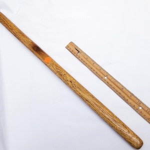 Peut inclure: Un long goujon en bois avec une extrémité arrondie et une pointe sombre, présentant un motif de grain de bois texturé. Un autocollant orange est apposé sur le goujon. Une règle en bois, marquée en pouces, est également présente.